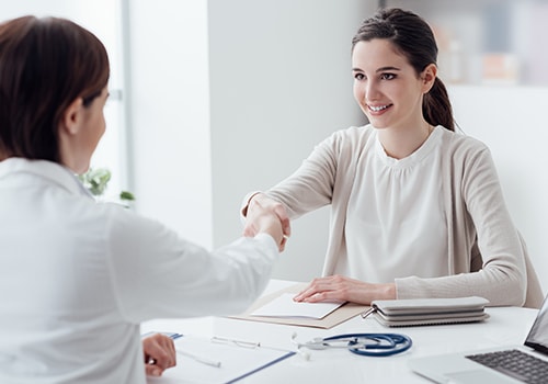 A clinician in a white lab coat, standing and speaking with a man and a woman, who are seated in a waiting room, with other clinicians in the background.