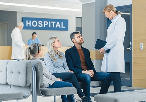 Two smiling women—a clinician and a patient—shaking hands across a desk topped with a stethoscope, papers and a laptop.