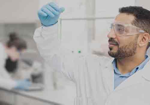 Male laboratory technician, wearing a lab coat, goggles and gloves, looking at a sample in his raised hand.